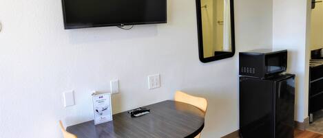 Una habitación pequeña con una mesa de madera oscura, dos sillas amarillas, un televisor negro montado en la pared, un microondas negro y un refrigerador.