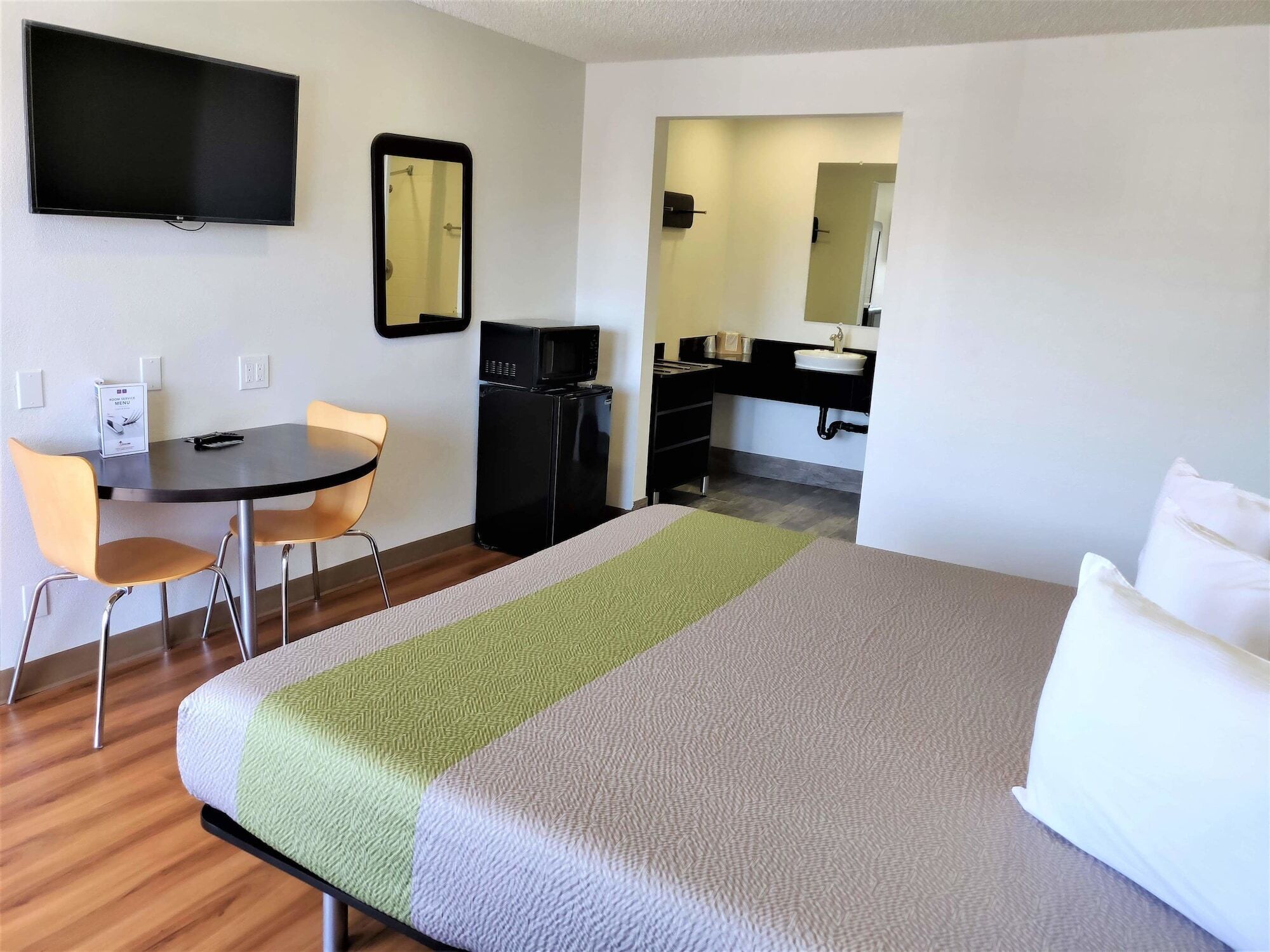 Habitación de hotel con cama, escritorio con silla, microondas, refrigerador pequeño, lavamanos y espejo.