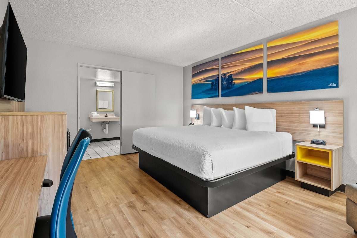 A modern hotel room with a bed, a desk, a chair, a TV, a bathroom, and a painting on the wall.