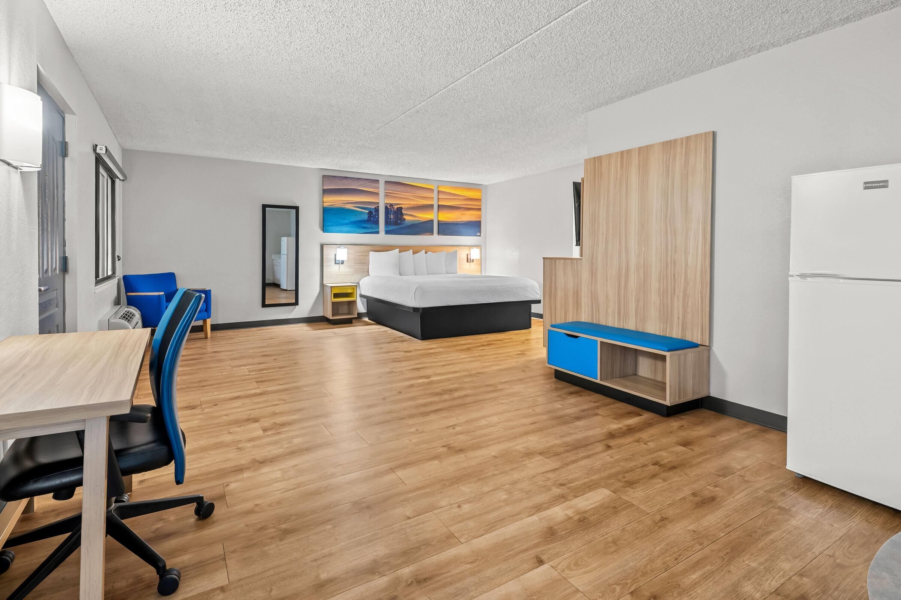 Una habitación de hotel moderna con suelo de madera, una cama con cabecero blanco, un escritorio con silla, un armario y un televisor fijado a la pared.