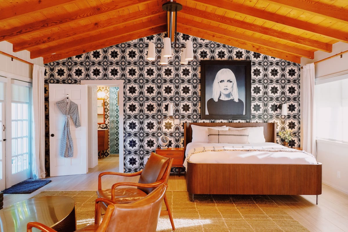 A bedroom with a large bed, a wooden ceiling, patterned wallpaper, and a hanging portrait.