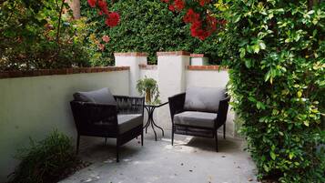 An outdoor patio area with two wicker chairs and a small table, surrounded by lush greenery and red flowers.