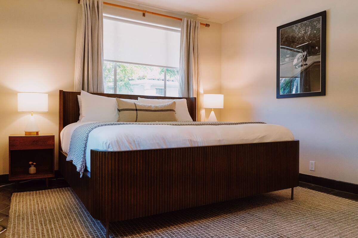 A neatly made bed with a wooden headboard, two bedside lamps, and a large window with curtains.