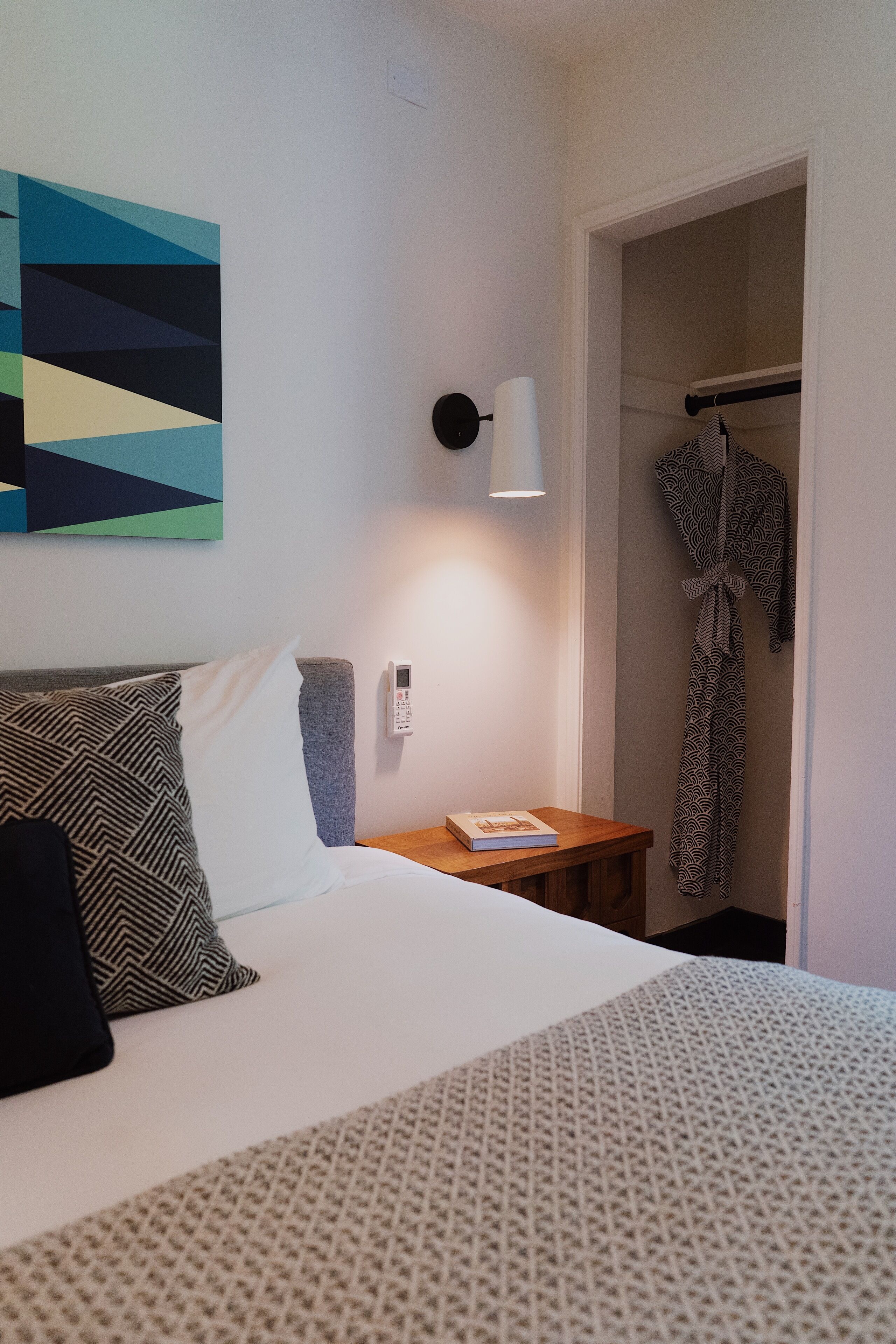 A hotel room with a bed, a bedside table, a lamp, a robe hanging on a rack, and a geometric artwork on the wall.