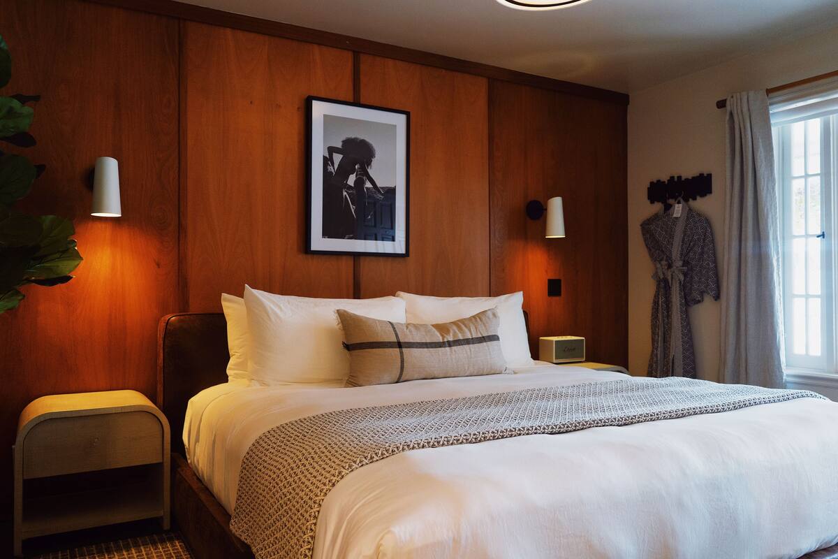 A neatly made bed with white linens and a patterned throw, a bedside table with a lamp, a framed photograph on the wall, and a window with curtains.
