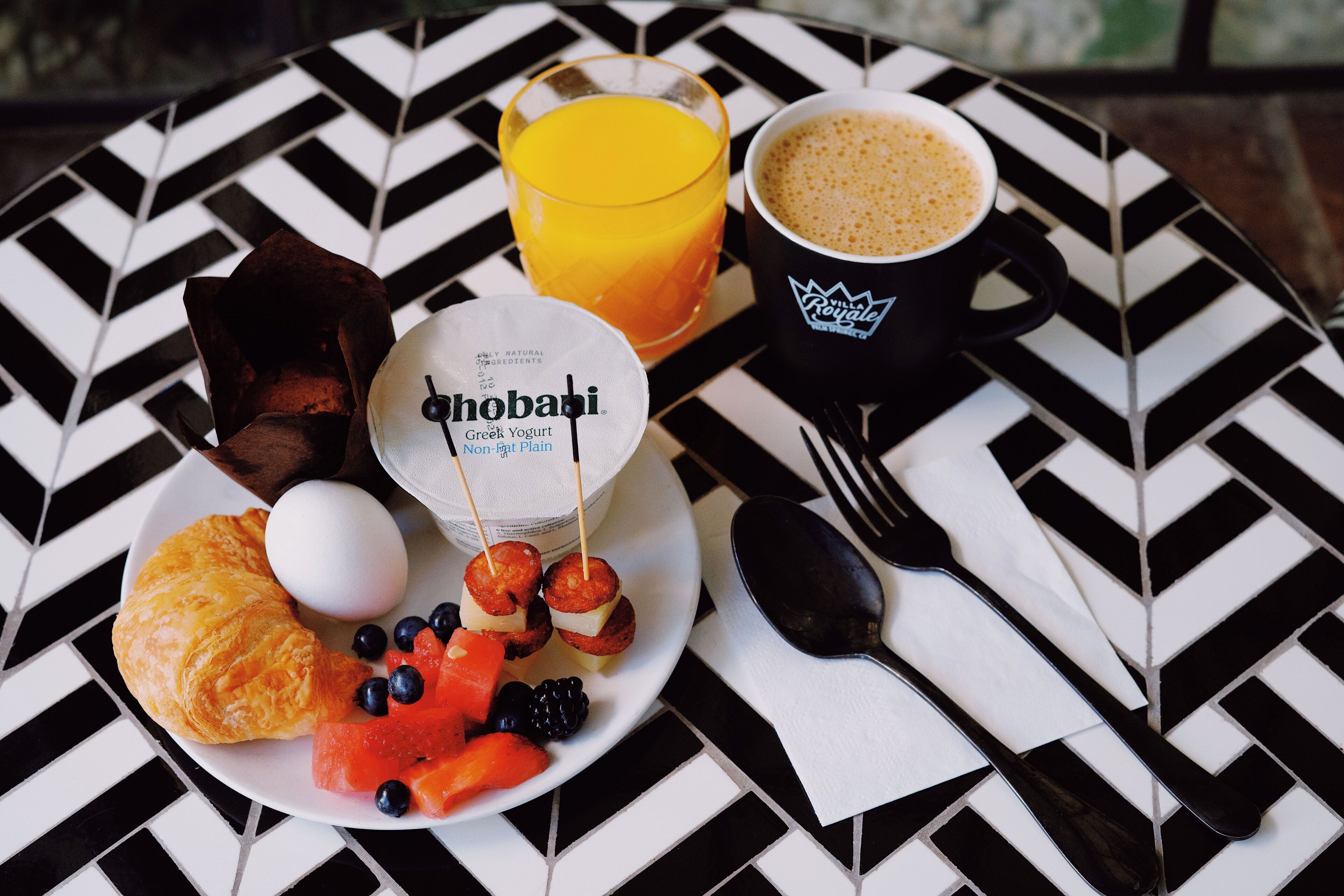 A breakfast plate with croissant, egg, blueberries, watermelon, and yogurt, a glass of orange juice, and a cup of coffee on a black and white patterned table.