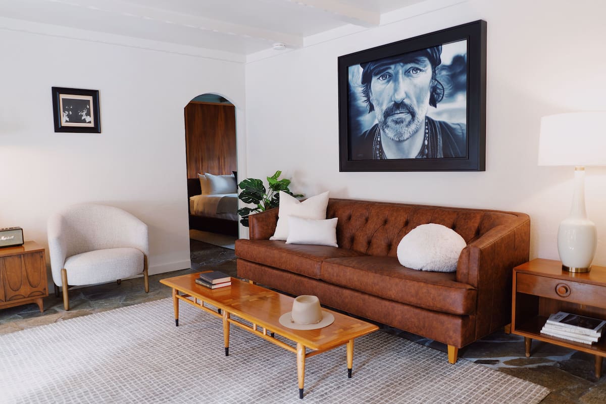 A living room with a brown sofa, a wooden coffee table, a white armchair, a side table with a lamp, and a framed artwork on the wall.