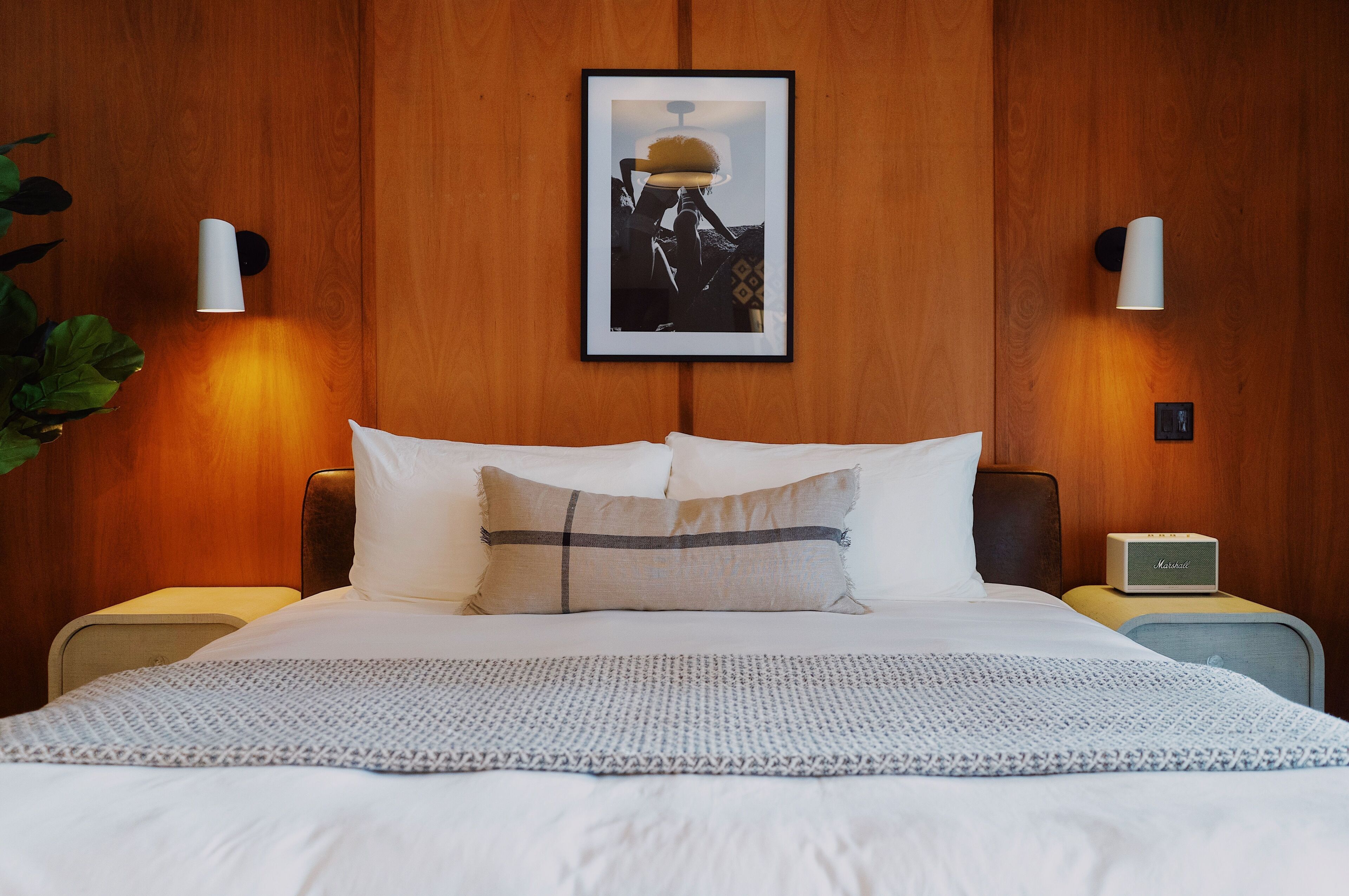A neatly made bed with a white duvet and a patterned throw, a framed picture on the wall, and a bedside table with a lamp and a radio.