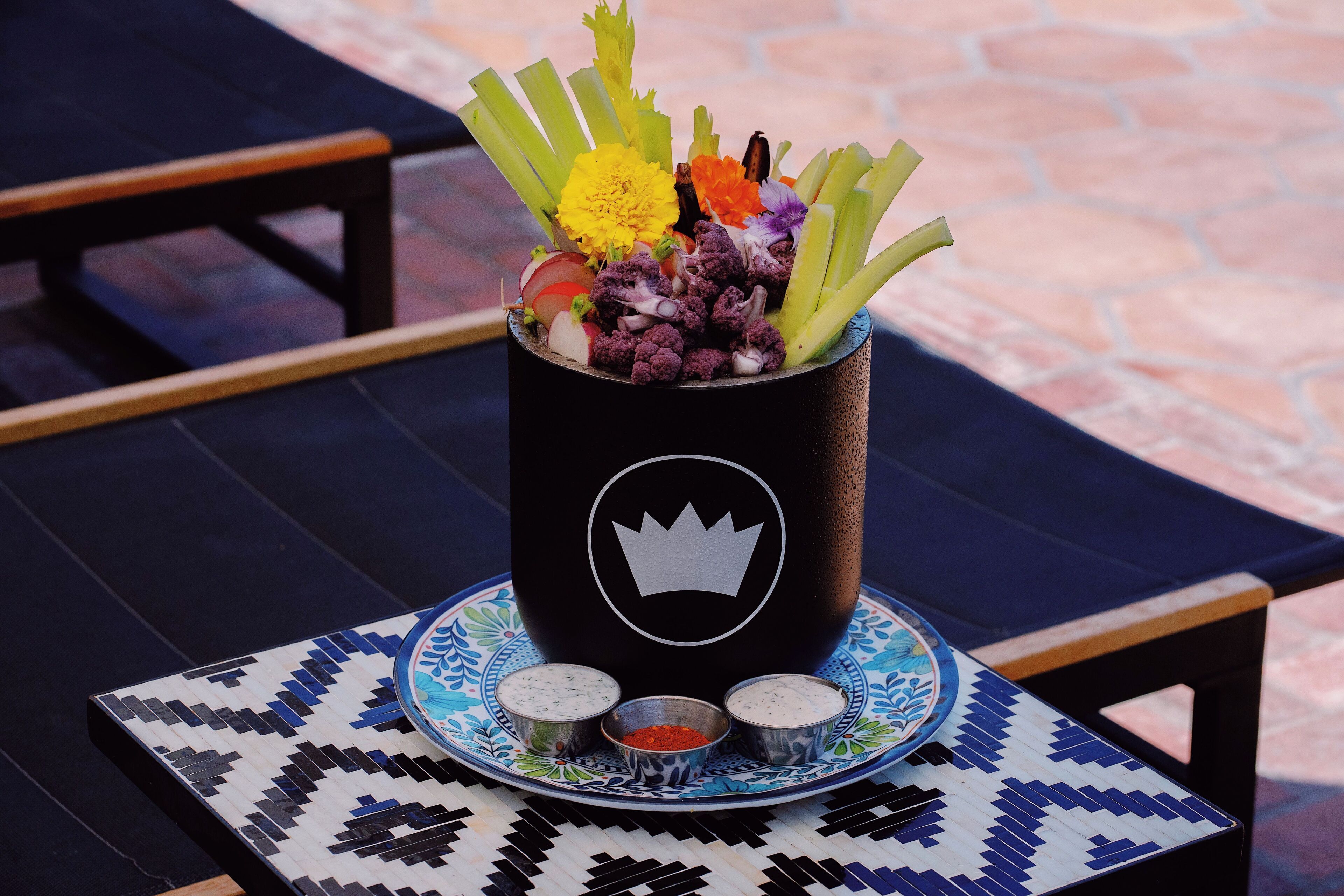 A beverage cup with a crown logo, filled with vegetables and a flower, placed on a patterned table with a plate and two small condiment containers.