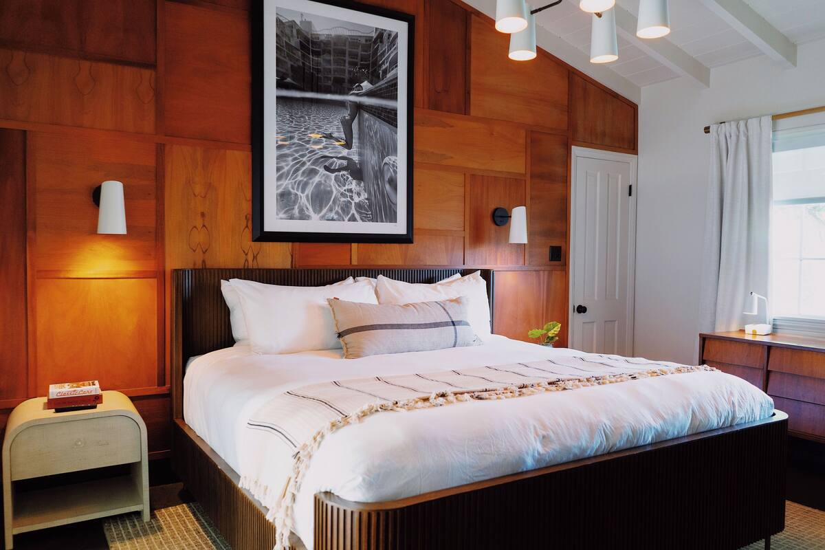 A neatly made bed with a striped blanket, a framed photograph above it, and a bedside table with a lamp.