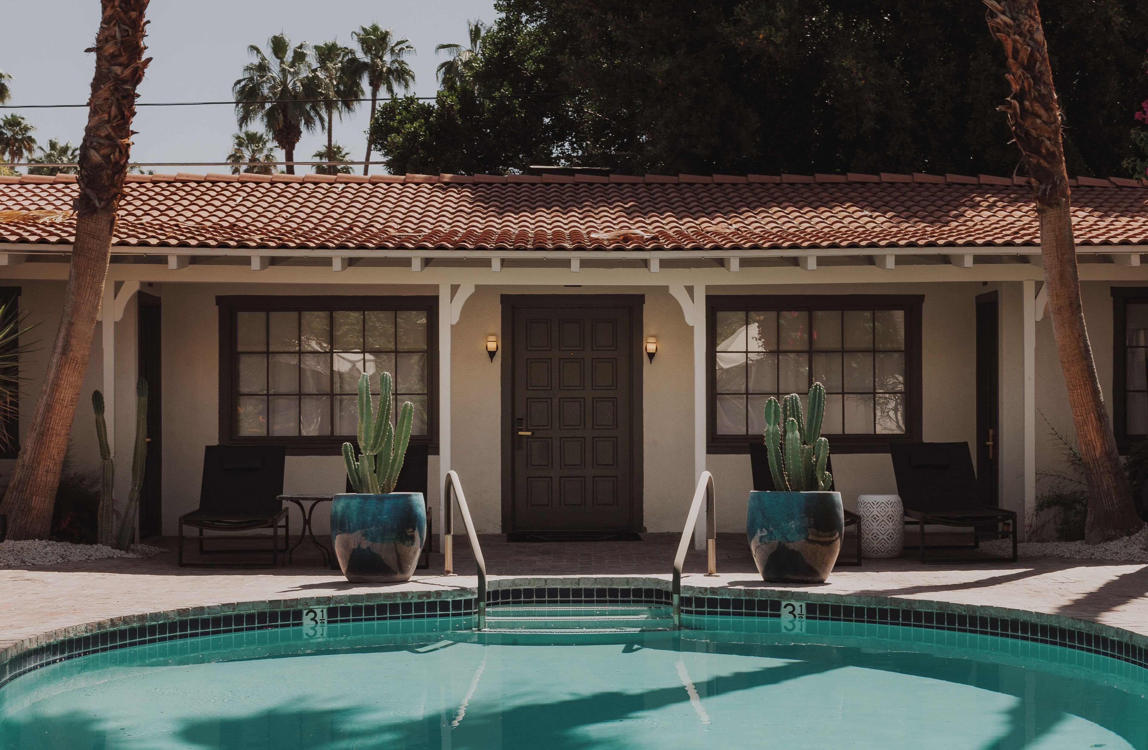 A pool area with a tiled deck, two potted cacti, and a building with a tiled roof and windows.
