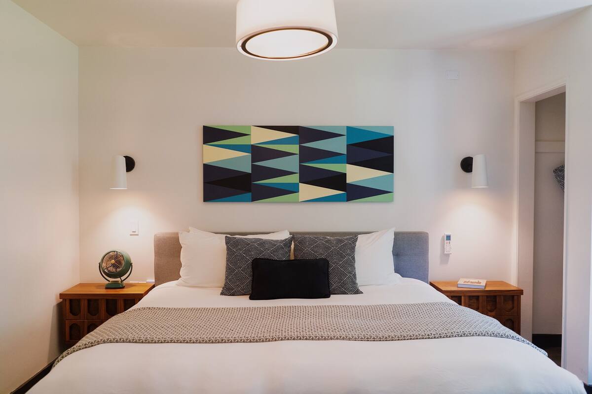 A neatly arranged hotel room with a bed, a bedside table with a lamp, a wall-mounted artwork, and a ceiling light fixture.