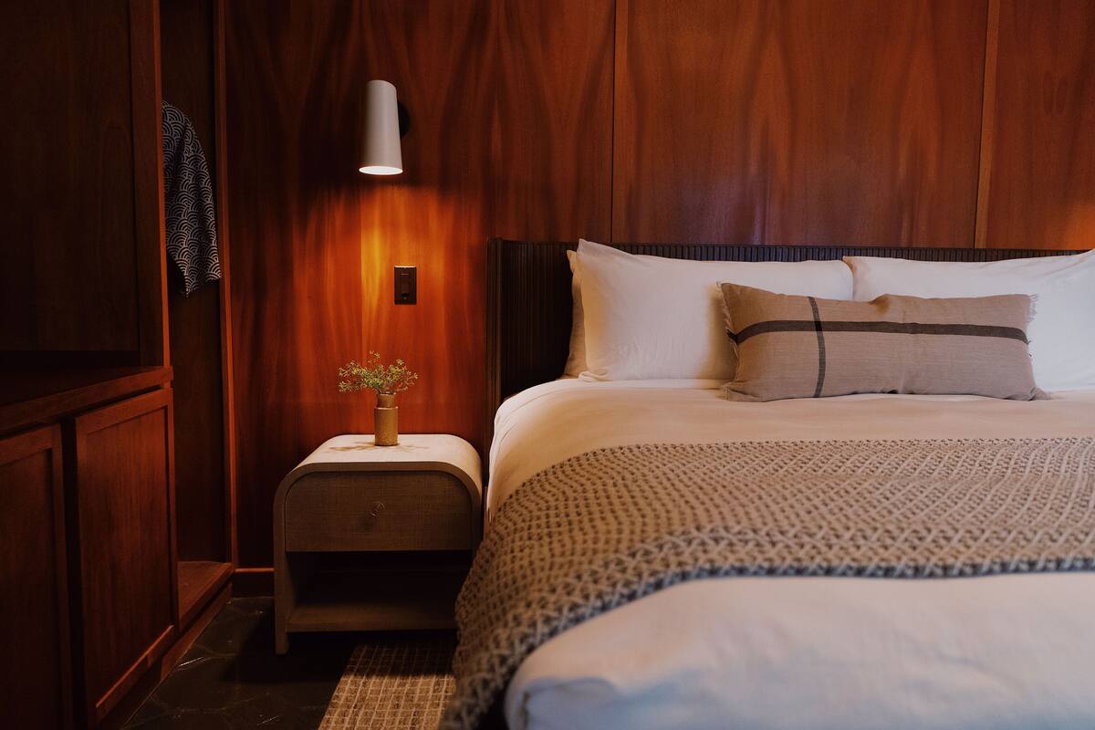 A hotel room with a wooden paneled wall, a bedside table with a vase of flowers, a bed with white bedding and a patterned throw, and a wall-mounted light fixture.