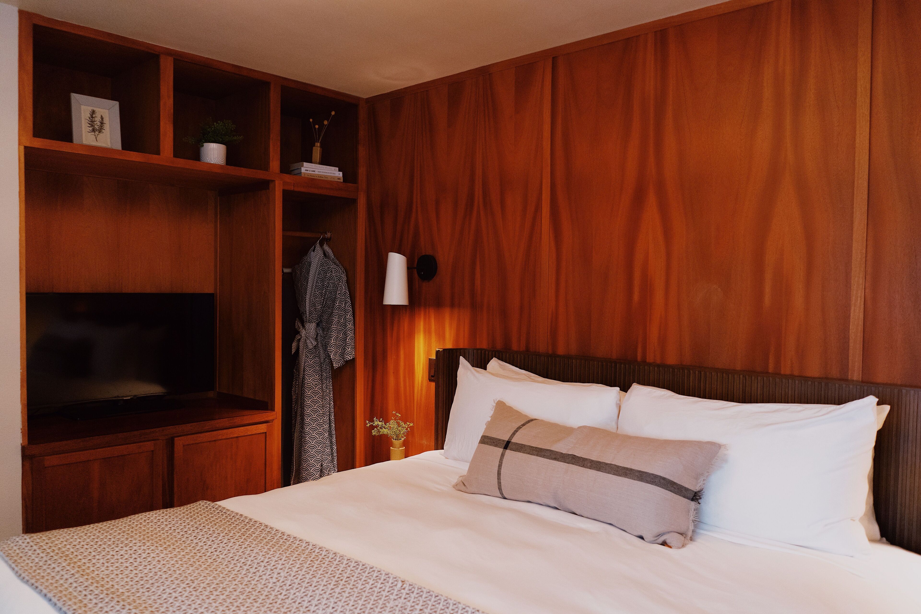 Une chambre d’hôtel avec une tête de lit en bois, un grand lit avec une literie blanche, un téléviseur posé sur une commode en bois et une petite table sur laquelle se trouve un vase de fleurs.