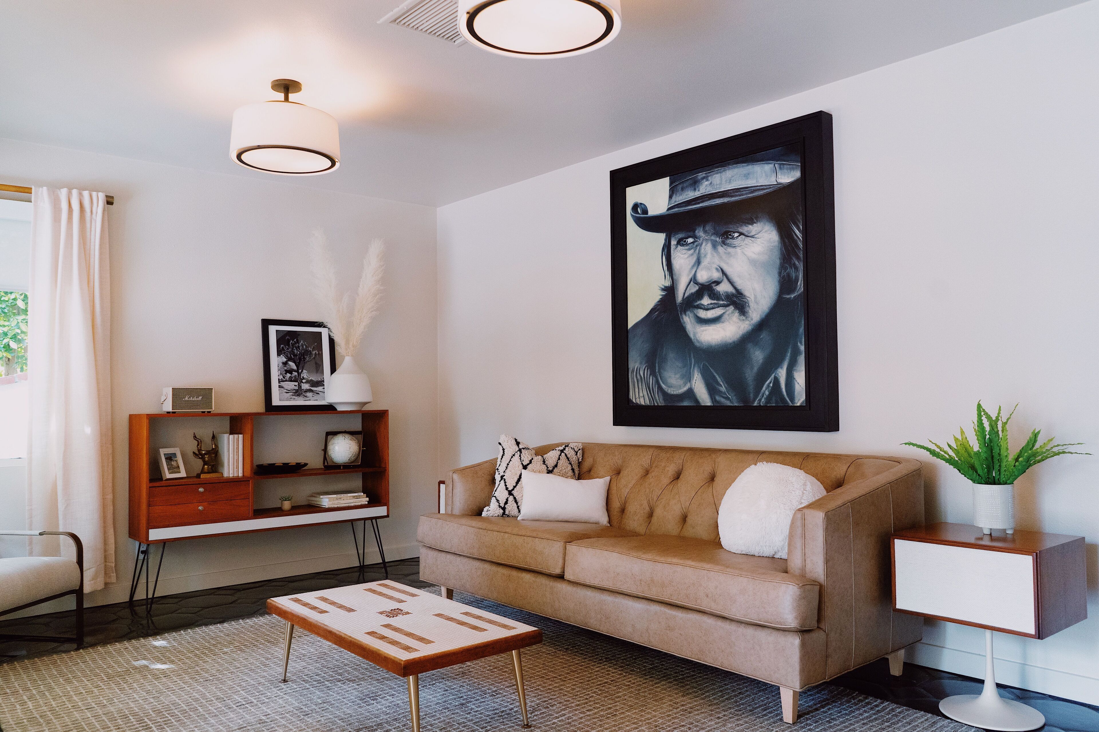 Een moderne woonkamer met een bruine leren bank, een houten bijzettafeltje, een witte fauteuil en een salontafel. Daarboven hangt een lijst met kunstwerk en op het bijzettafeltje staat een potplant.