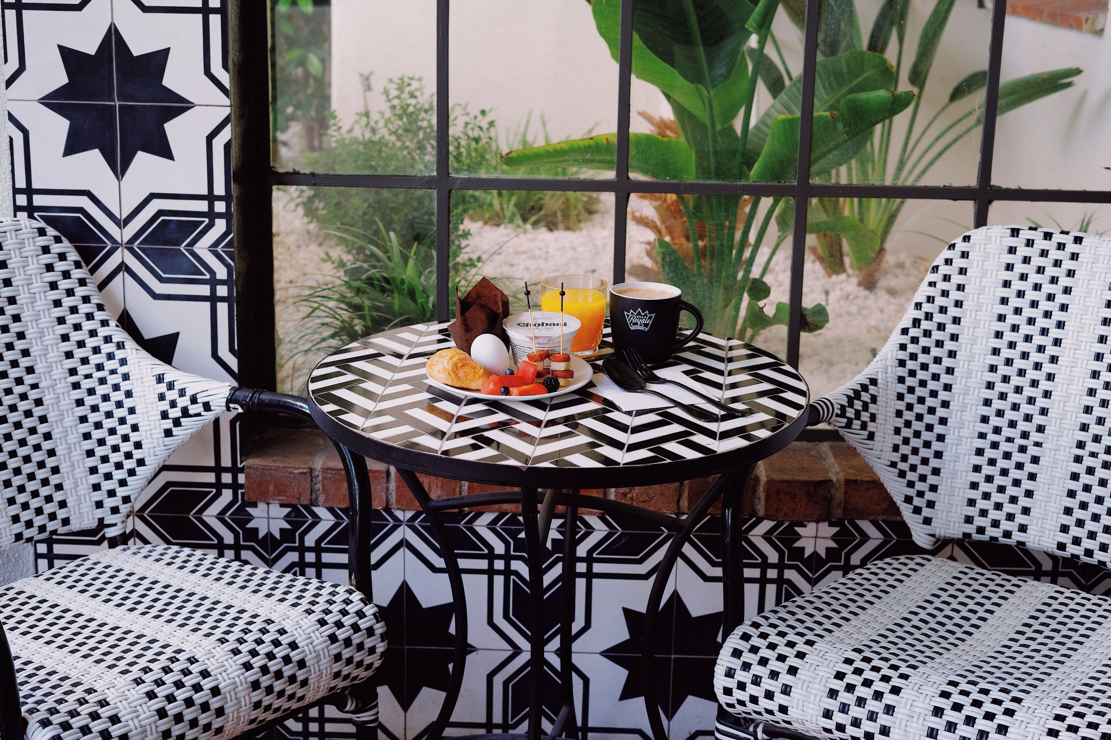 Une table recouverte d’une nappe à motifs, deux chaises au design géométrique et une table dressée pour le petit-déjeuner avec des œufs, des toasts, du jus de fruits et une tasse de café.