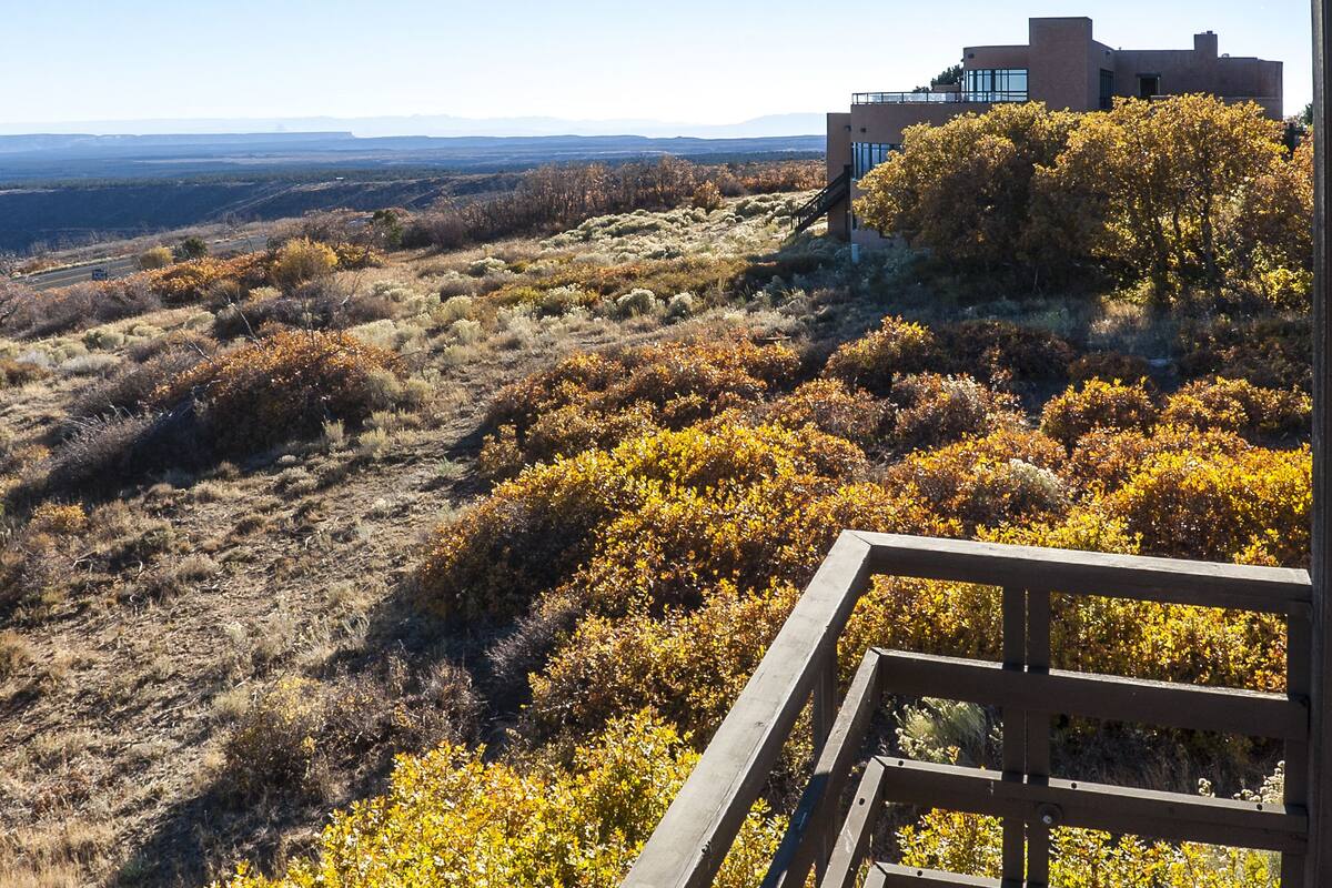 Una terraza de madera con vistas a un paisaje montañoso, follaje otoñal, un edificio moderno con amplios ventanales y cielo despejado.