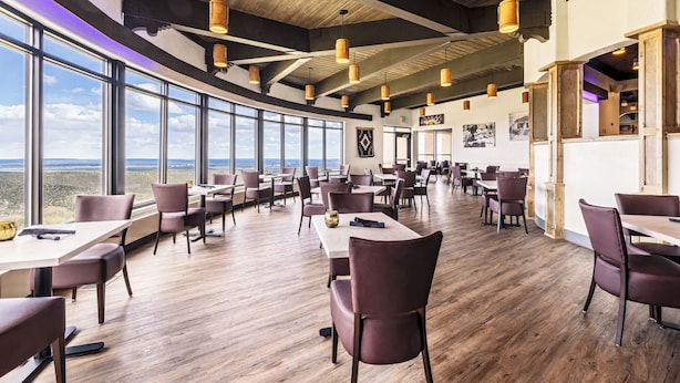 A spacious dining area with high ceilings and large windows offering a view of the landscape. Tables and chairs are arranged for dining.