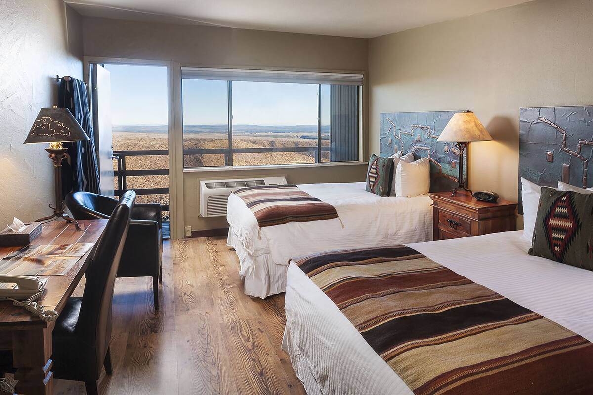 Habitación de hotel con dos camas, un escritorio con teléfono, una silla, una lámpara y vistas al desierto a través de ventanas amplias.