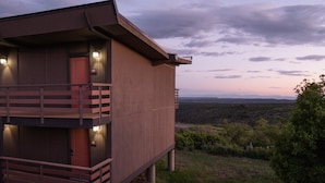 A two-story wooden cabin with a balcony, situated on a grassy hill with a view of the landscape.