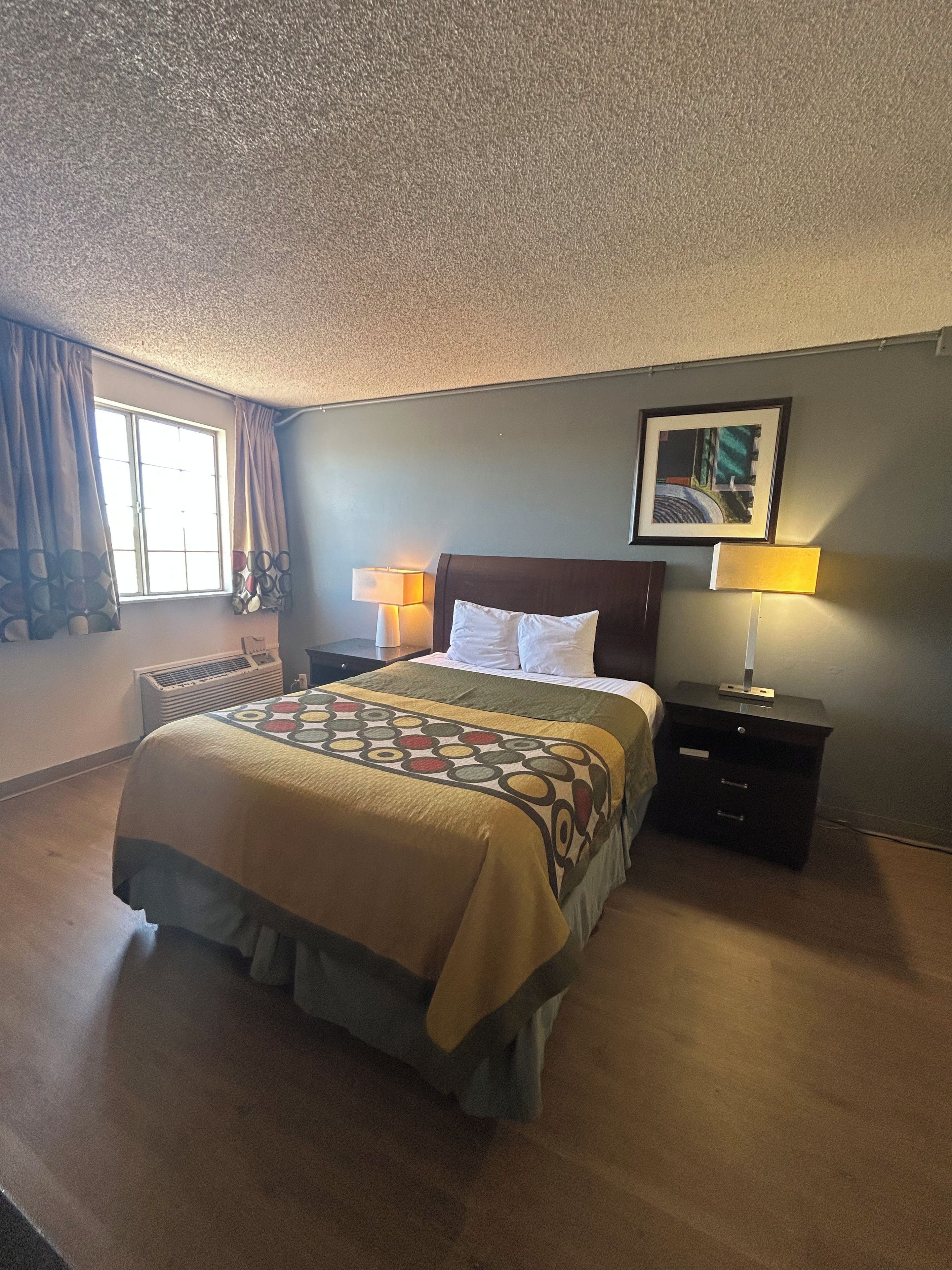Una habitación de hotel con una cama, dos mesitas de noche con lámparas, una ventana con cortinas y un cuadro enmarcado en la pared.