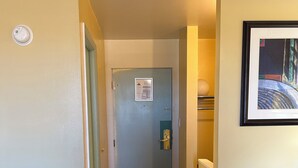 Un pasillo estrecho con suelo de madera, una puerta con cerradura de seguridad, una lámpara fijada a la pared y un cuadro enmarcado.