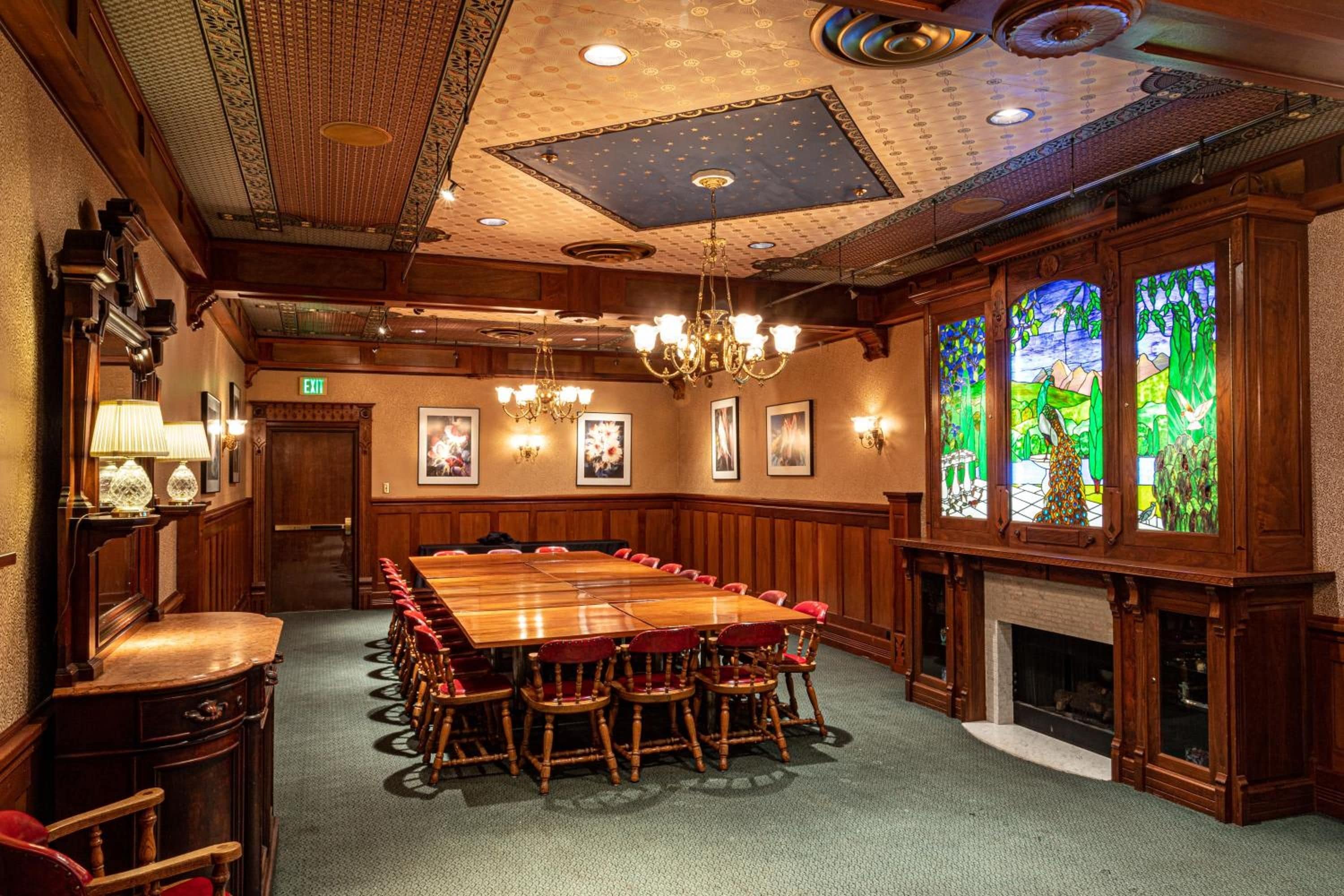 A conference room with a large wooden table, red chairs, chandeliers, framed pictures on the walls, and stained glass windows.