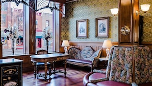 Une pièce avec un plancher en bois, une table ronde, un banc et un fauteuil à motifs floraux. Des tableaux encadrés ornent le mur, et une lampe se trouve sur une table d’appoint.