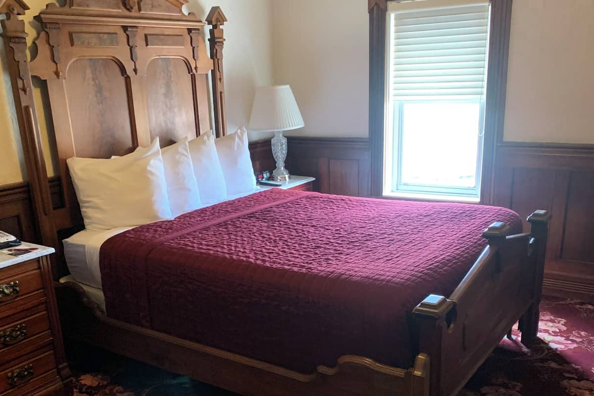 A bedroom with a large bed, a wooden headboard, a nightstand with a lamp, and a window with blinds.