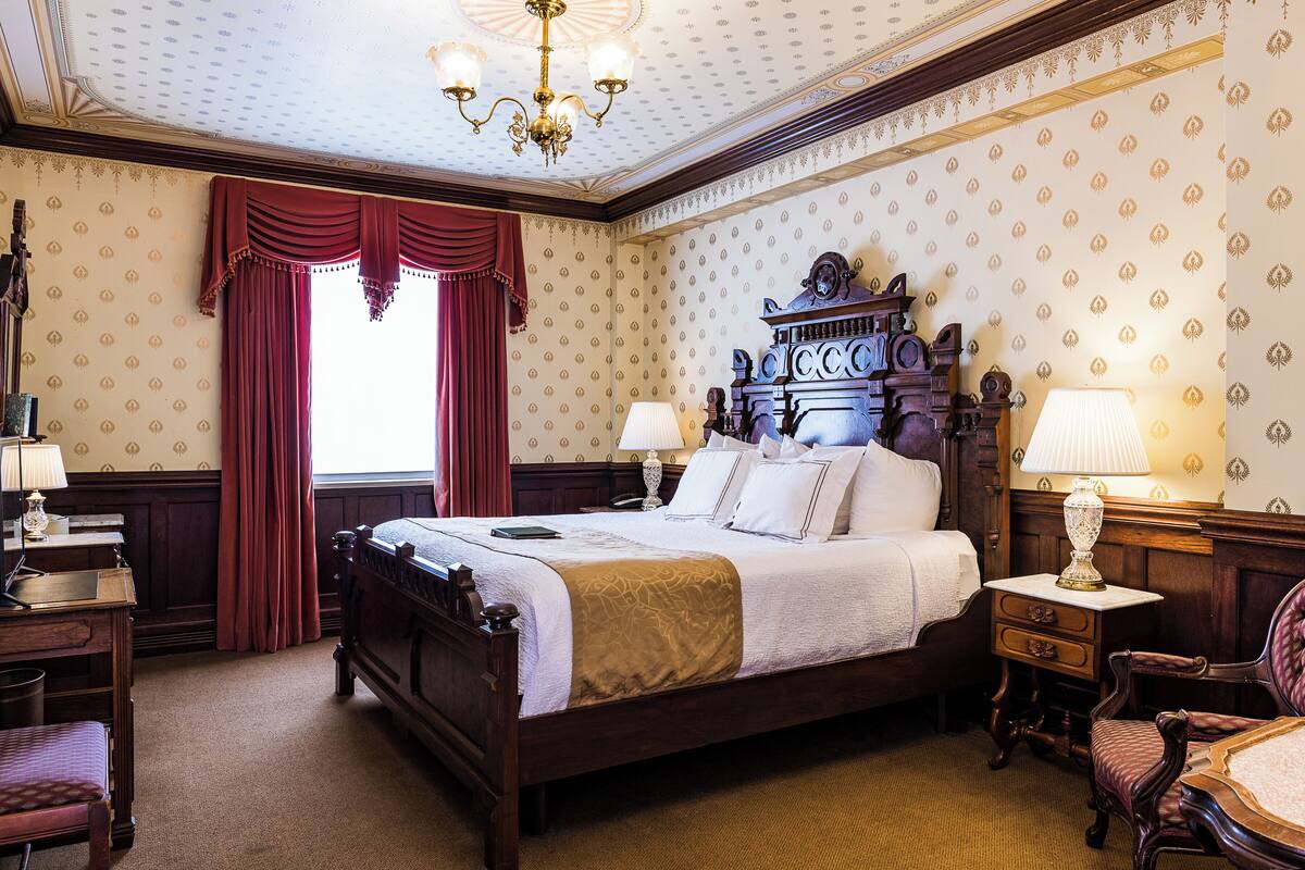 A classic bedroom with a four-poster bed, two bedside tables with lamps, a wooden chair, and a large window with red curtains.