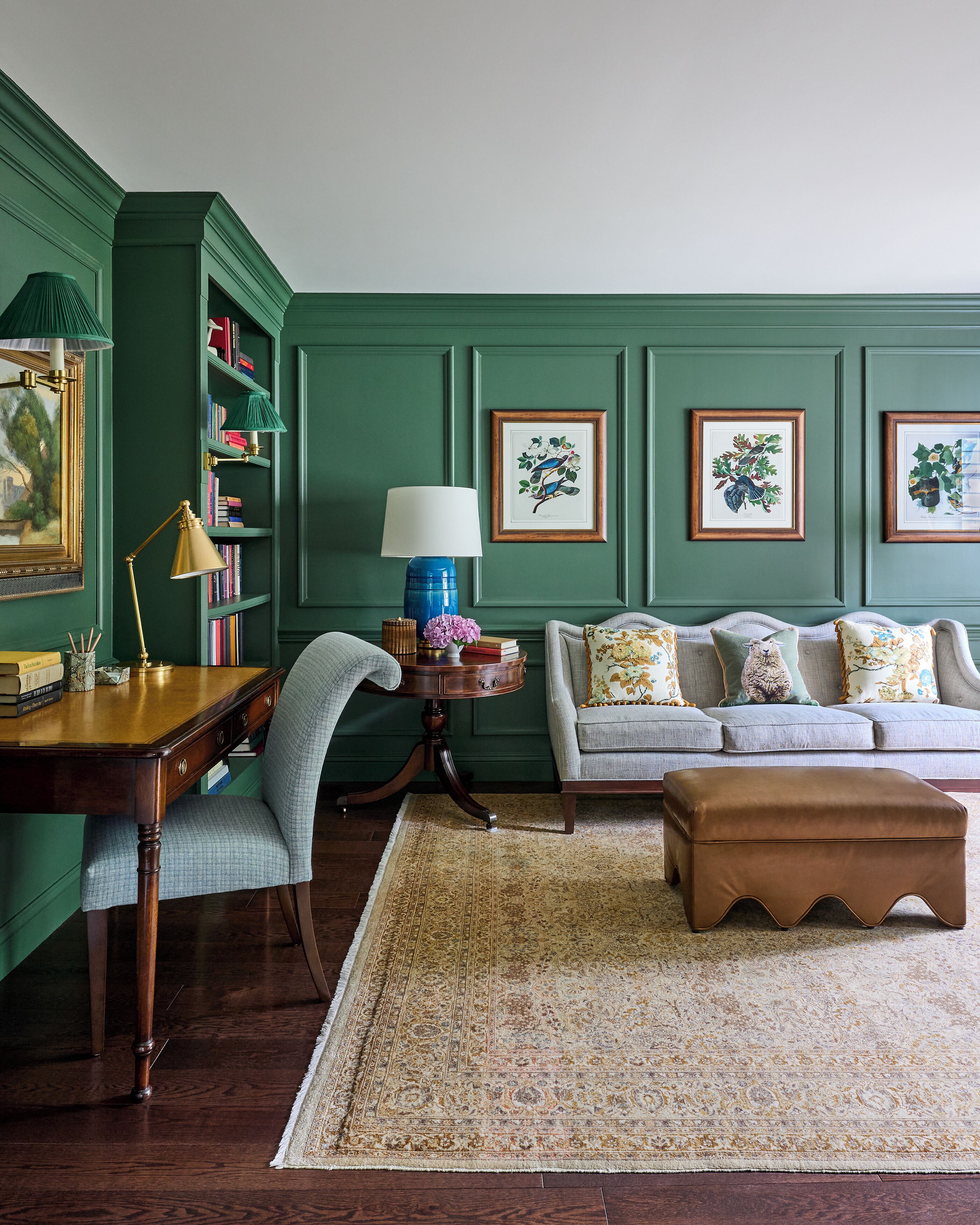 A room with a wooden desk, a blue lamp, a green sofa with patterned cushions, and framed botanical prints on the wall.