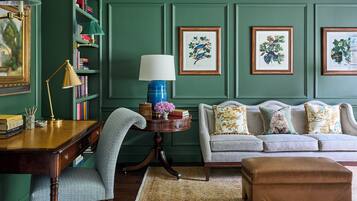 A room with a wooden desk, a blue lamp, a green sofa with patterned cushions, and framed botanical prints on the wall.