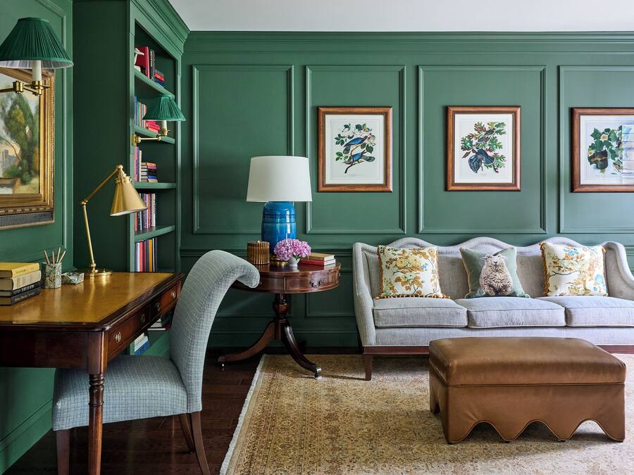 A room with a wooden desk, a blue lamp, a green sofa with patterned cushions, and framed botanical prints on the wall.