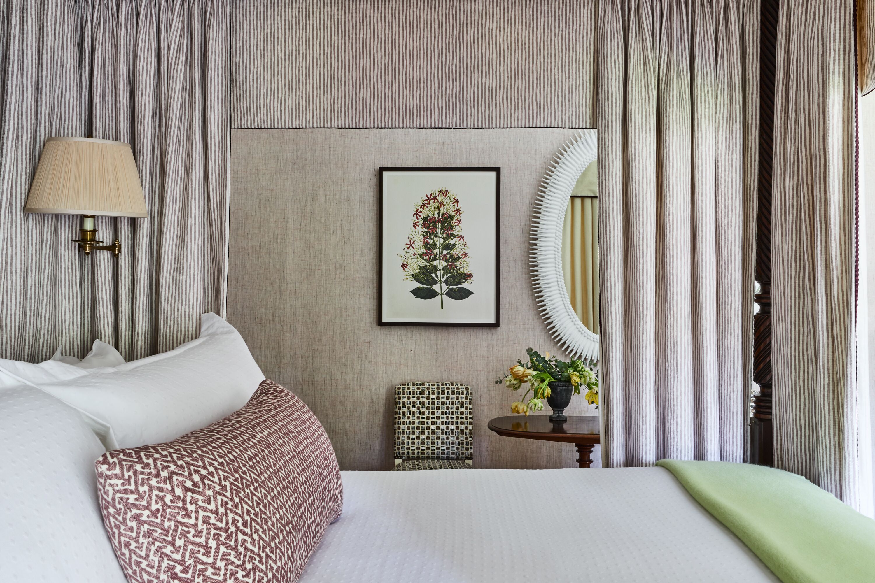 A neatly arranged bedroom with a bed, a bedside lamp, a framed botanical print, a small table with flowers, and a chair.