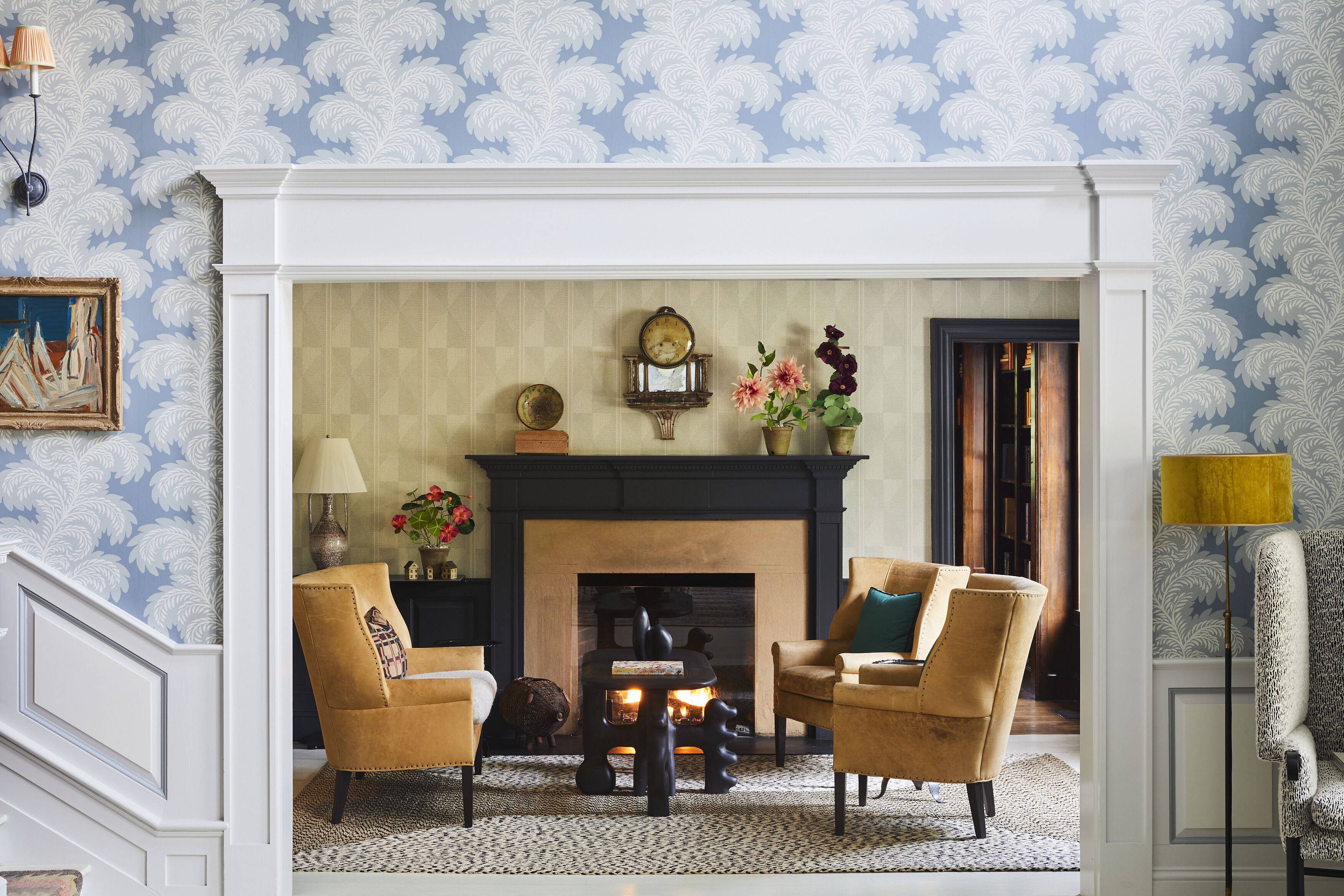 A room with a fireplace, two armchairs, a lamp, and a patterned wallpaper.