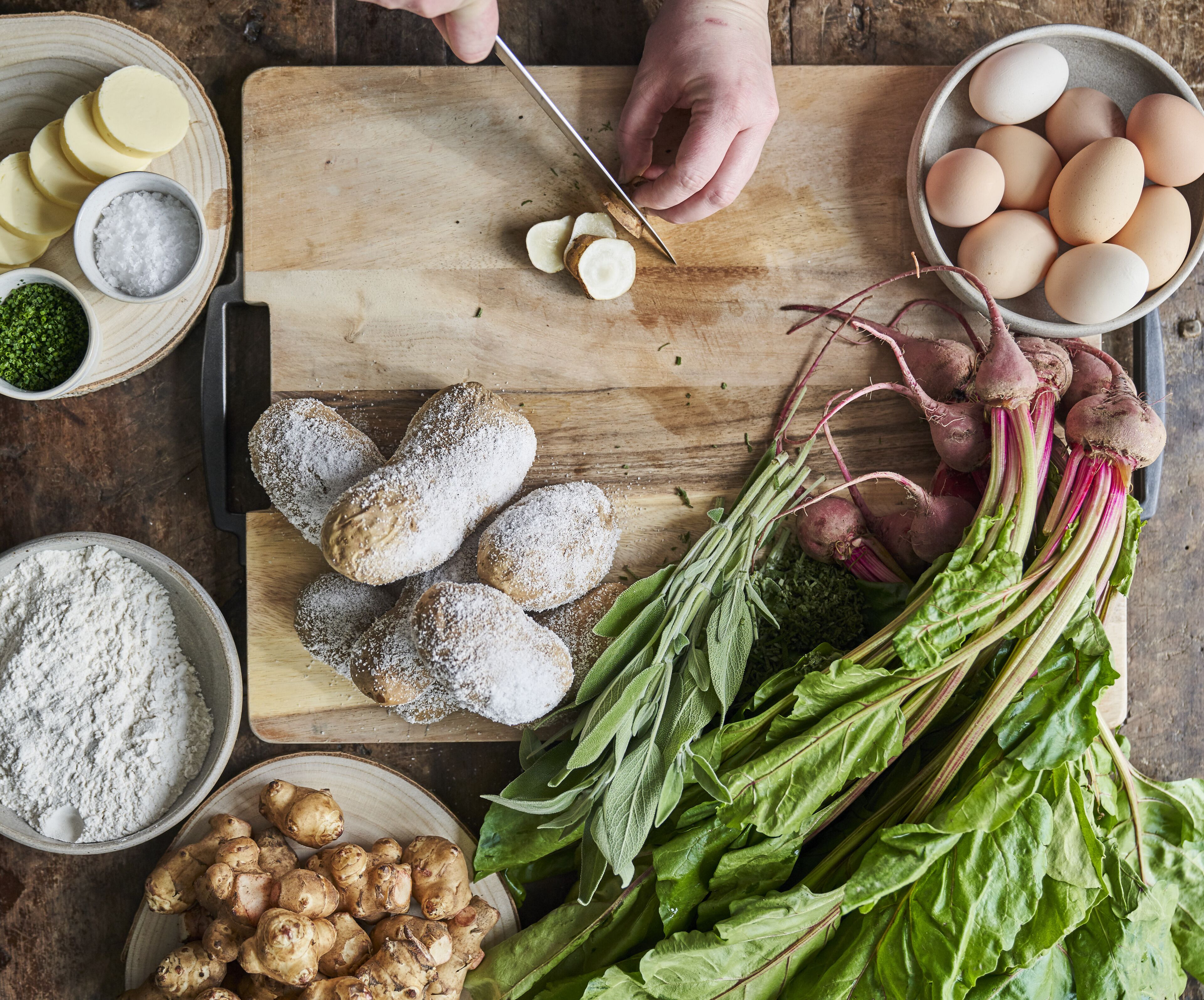 A wooden cutting board with sliced potatoes, a bowl of eggs, and various vegetables like beets and spinach.