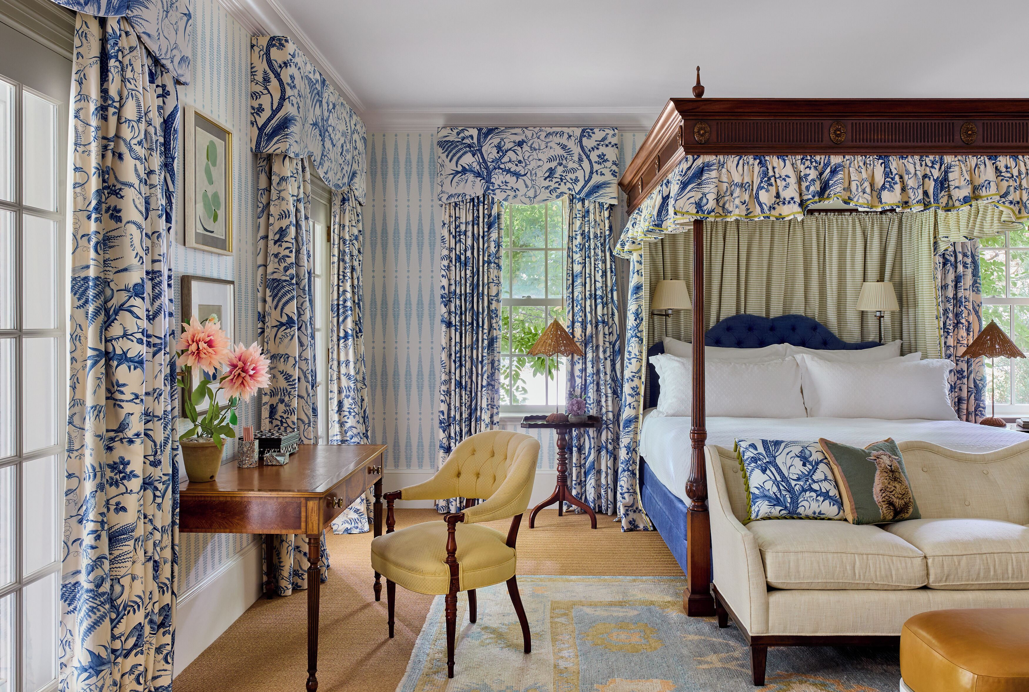 A bedroom with a four-poster bed, a sofa, a chair, and a desk with a vase of flowers.