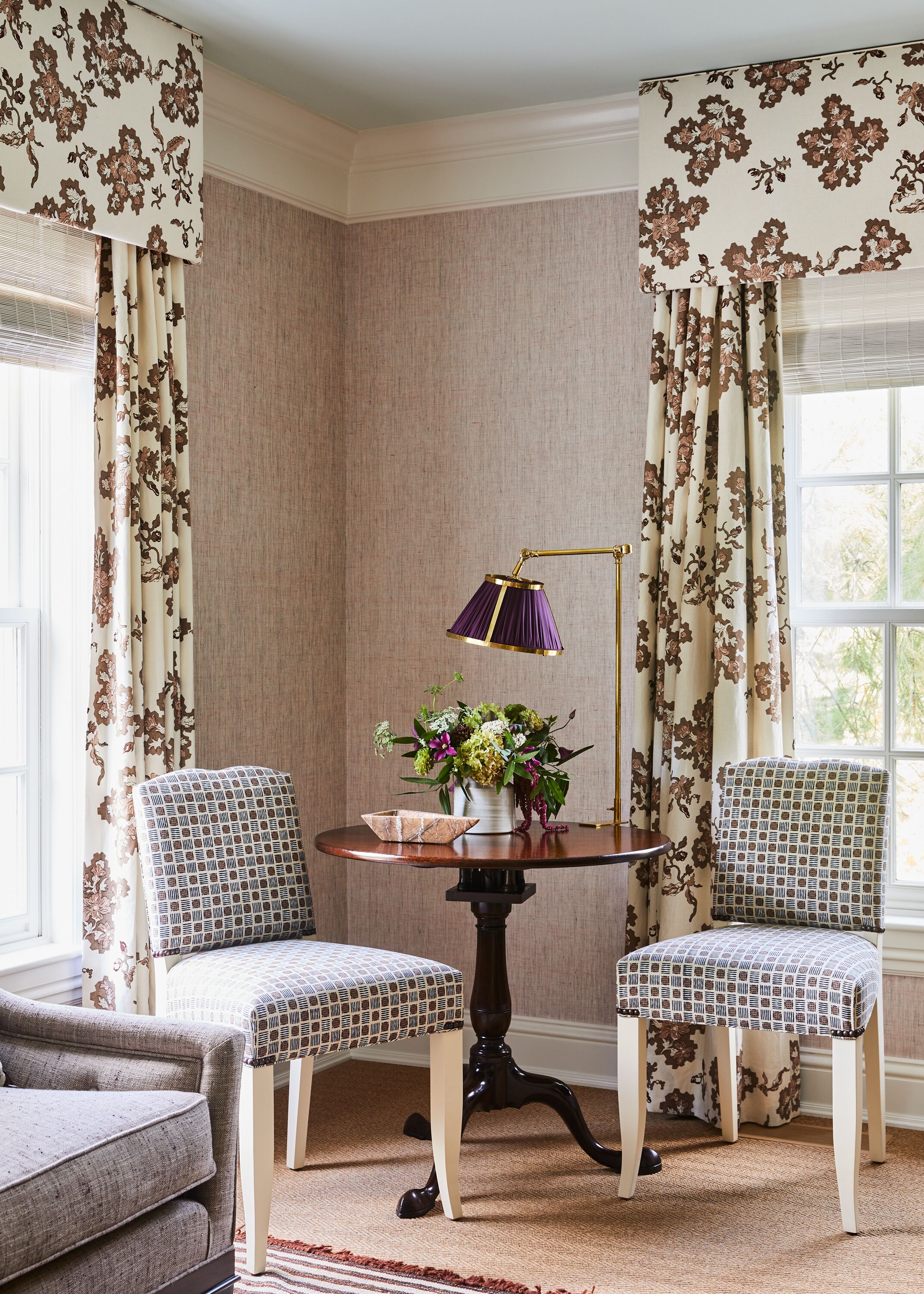 A corner room with two patterned chairs, a round table with a lamp, and a floral arrangement.