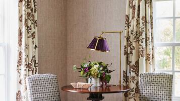 A corner room with two patterned chairs, a round table with a lamp, and a floral arrangement.