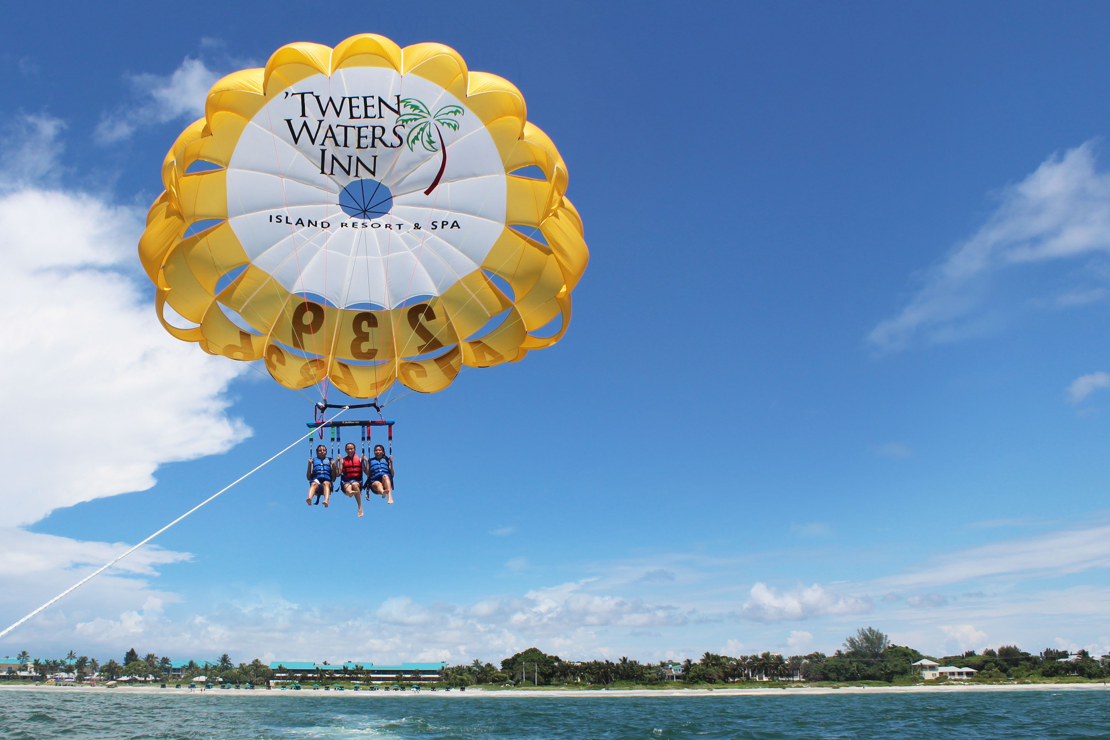 A parasail with 'TWEEN WATER'S INN' branding, over a beach with clear skies.