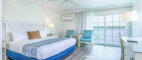 A hotel room with a bed, two chairs, a desk, a ceiling fan, and a view of the outdoors.