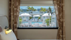Habitación de hotel con cama, ventana con cortinas y vista a un área de piscina con sombrillas y sillas de descanso.