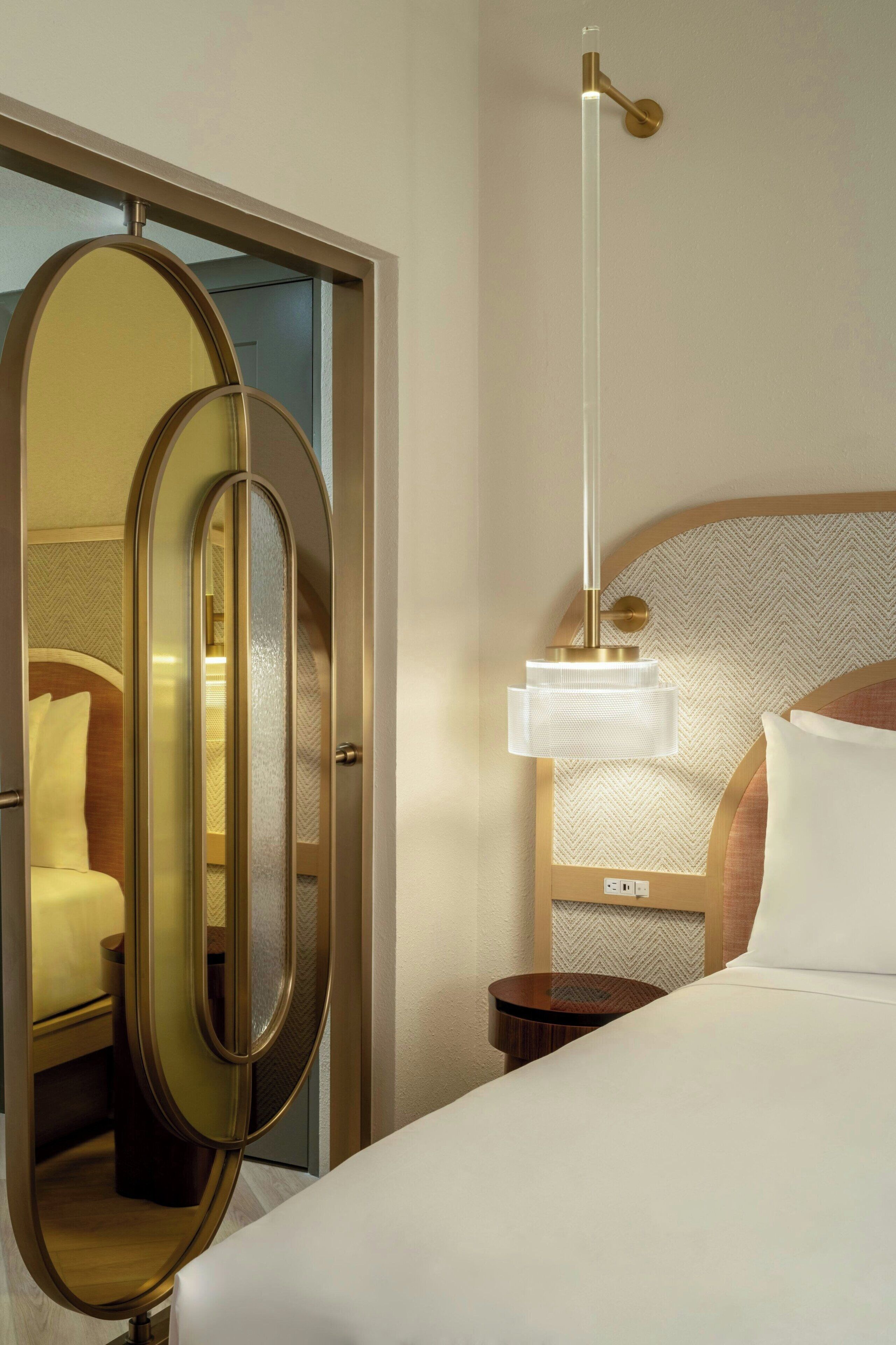 Una habitación de hotel con cama, mesita de noche, espejo y lámpara colgante.