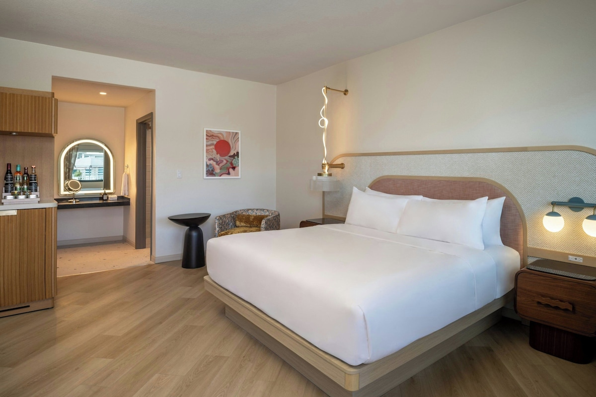 A modern hotel room with a large bed, bedside tables, a desk with a mirror, and a small seating area.