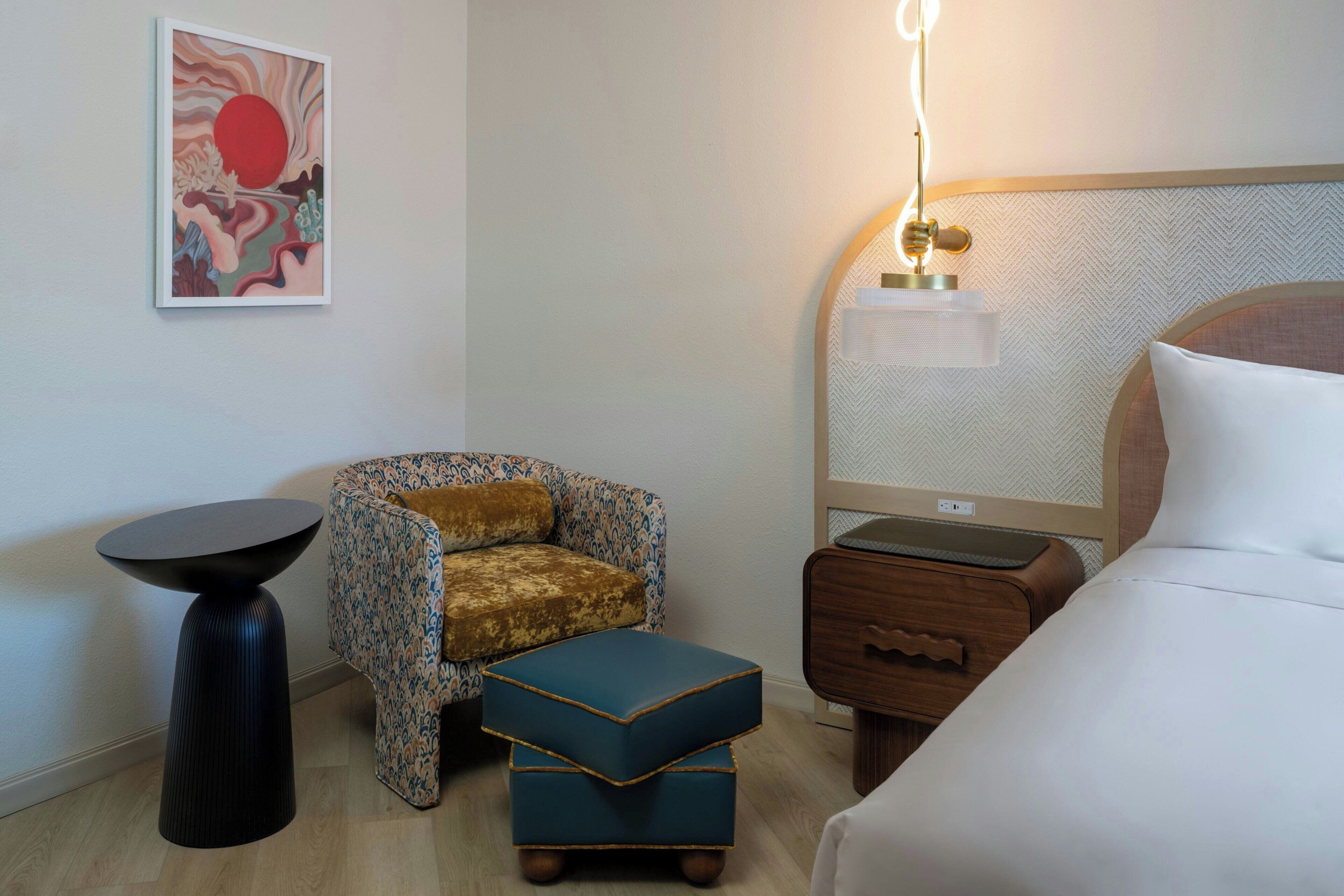 Una habitación de hotel con una cama, una mesita redonda pequeña, un sillón estampado, un reposapiés azul, una mesita de noche y una lámpara fijada a la pared.