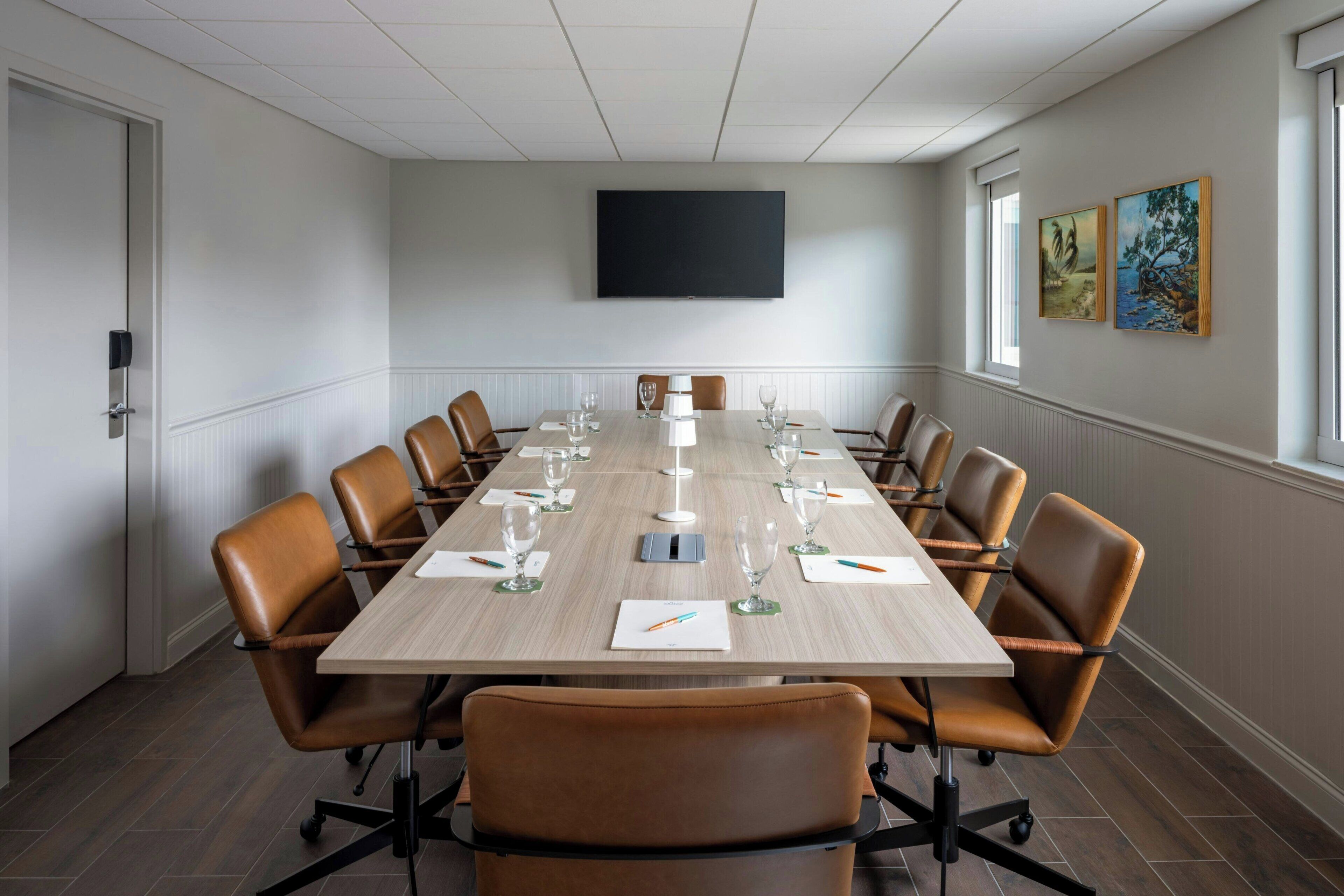 A conference room with a long table, brown leather chairs, a flat-screen TV on the wall, and artwork on the wall.