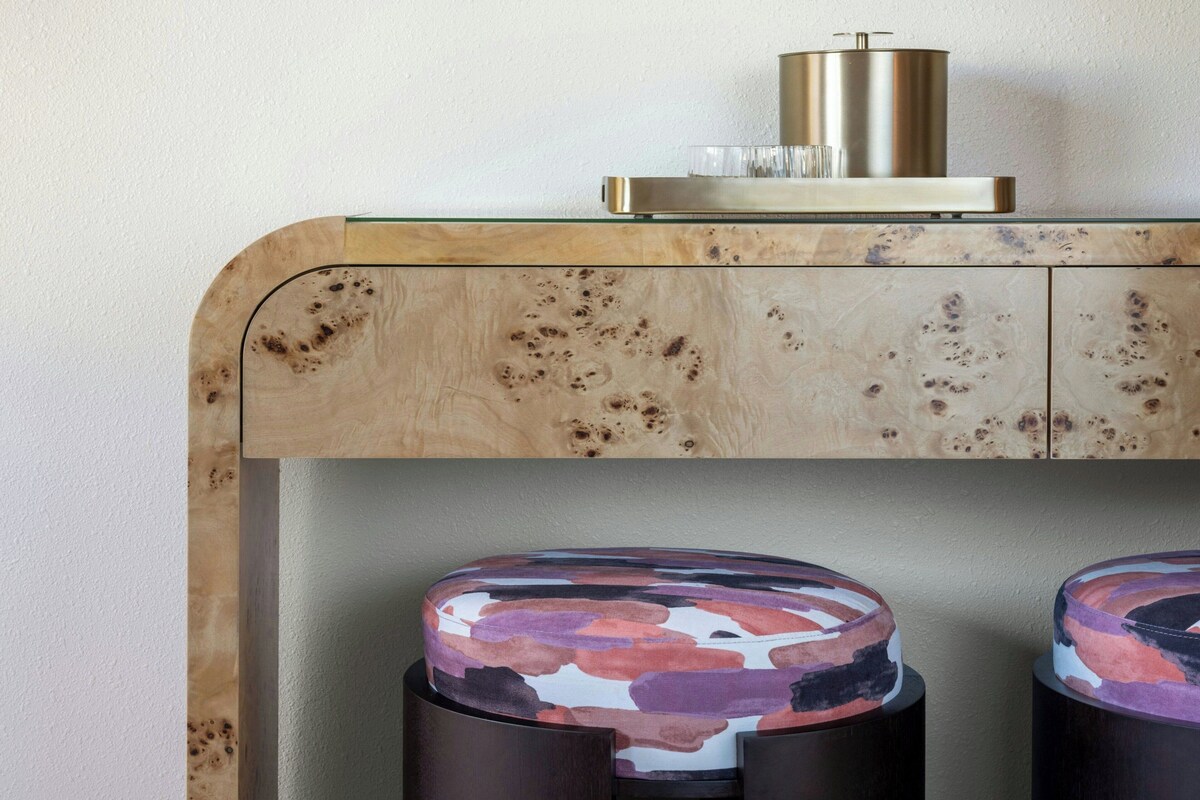 A wooden console table with a glass top, a metallic container, and two patterned stools.