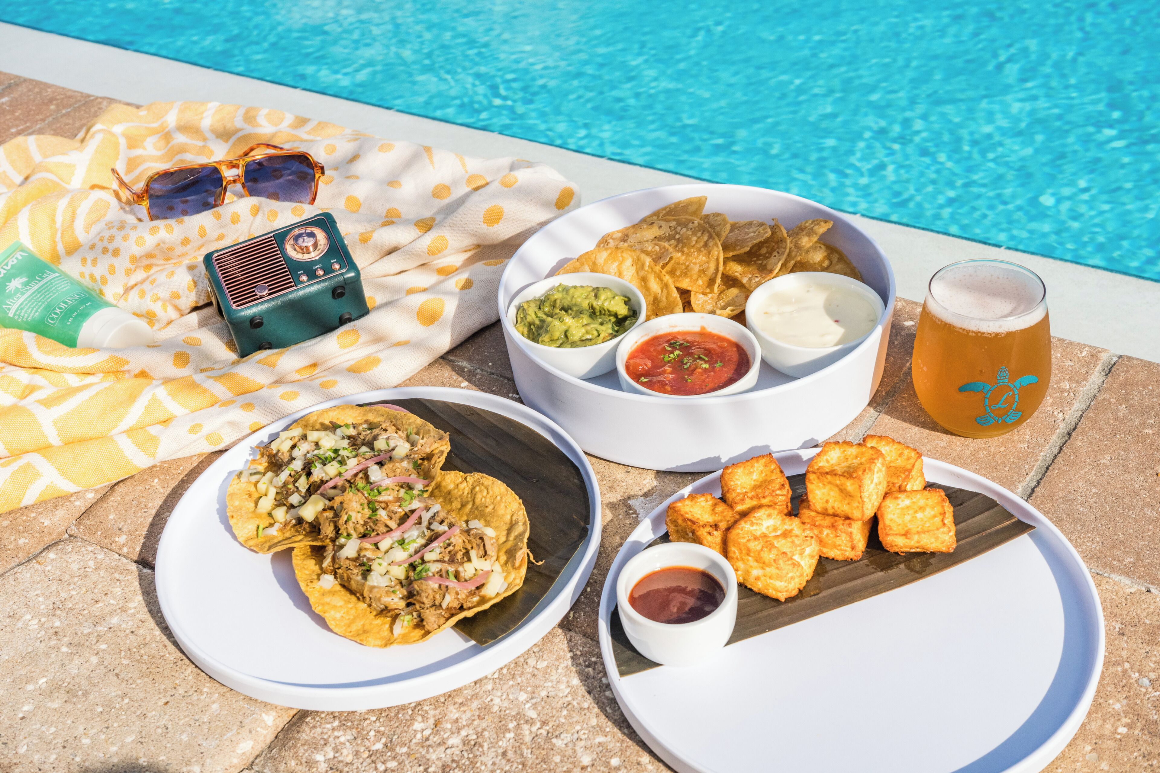 A poolside setting with a table set for a meal including tacos, chips, and beverages.
