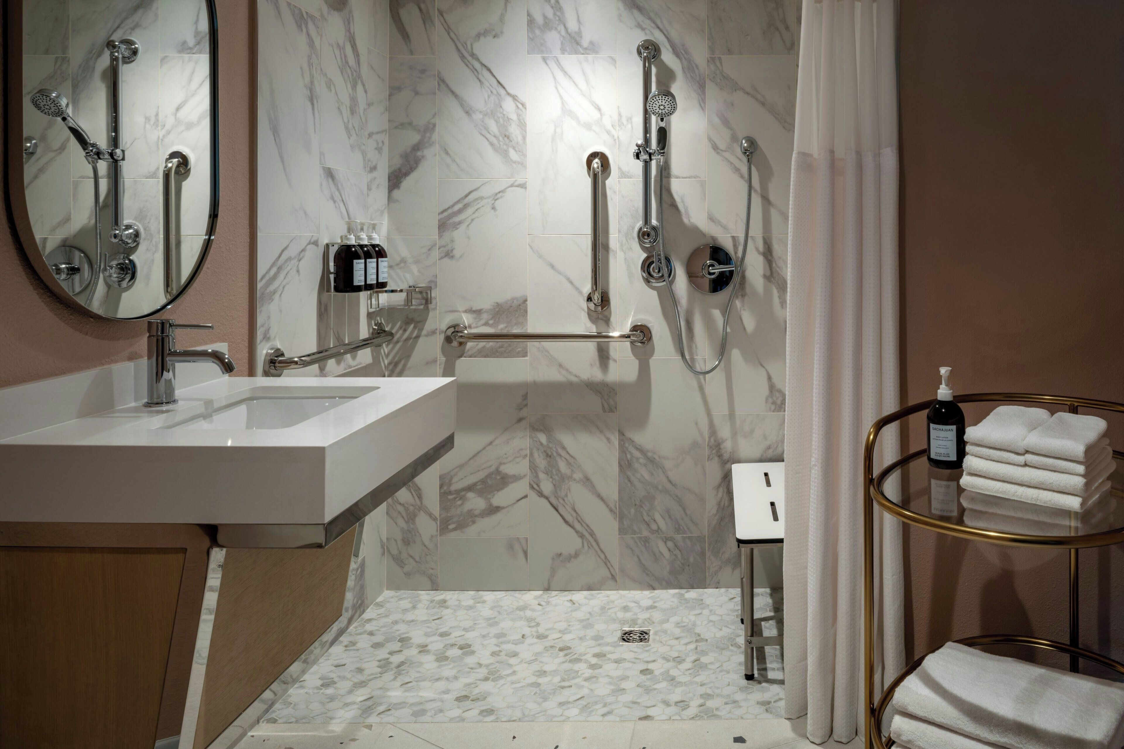 A bathroom with a sink, mirror, shower with grab bars, and a towel rack.
