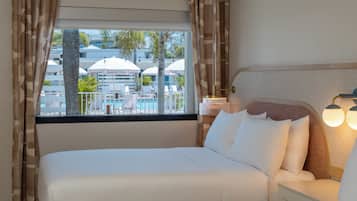 A hotel room with a large bed, a nightstand, a lamp, and a view of a pool area.